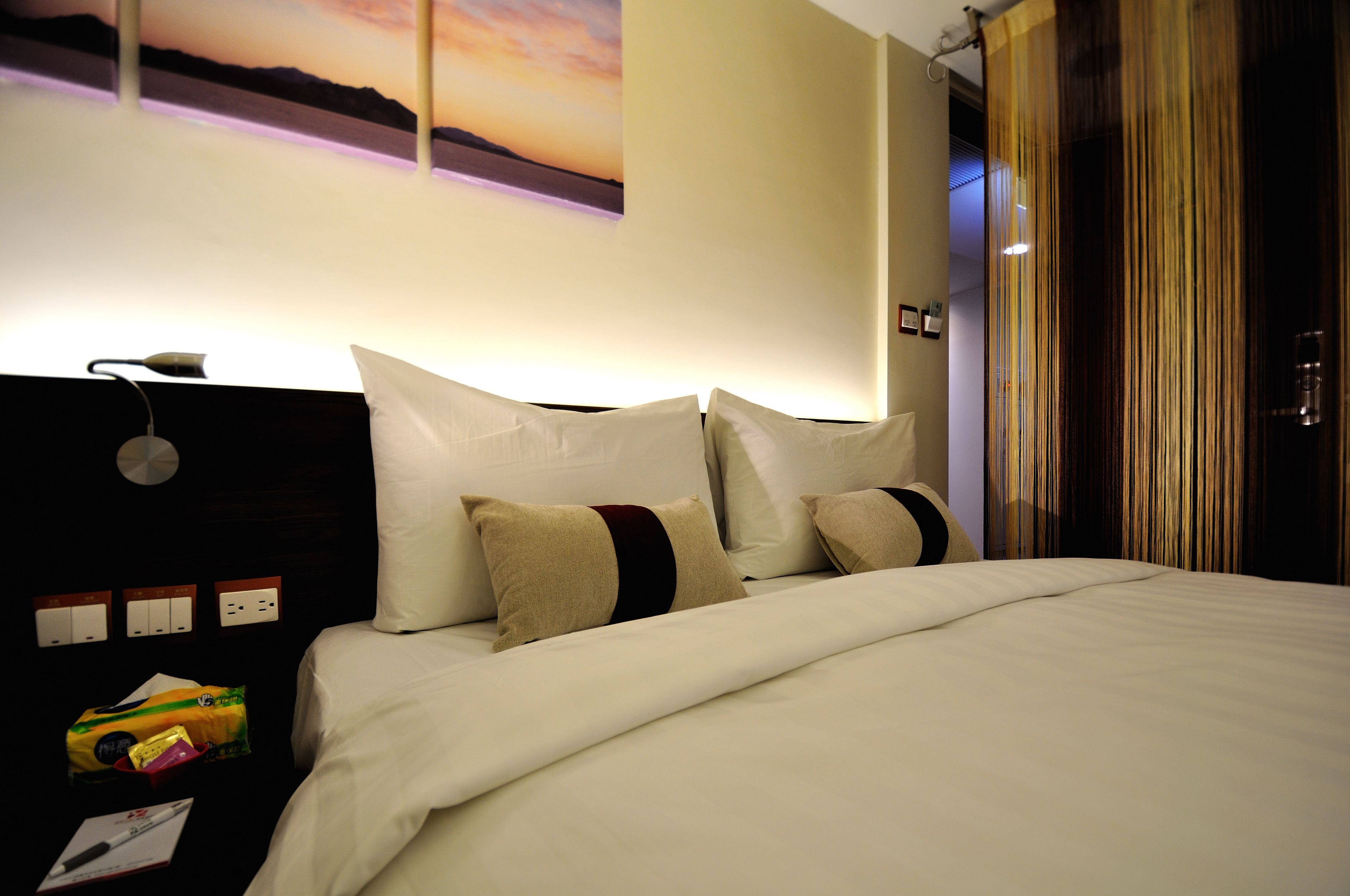 A neatly made bed with white bedding and multiple pillows, a wall-mounted lamp, and a painting of a sunset.