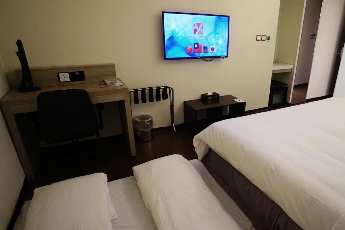 A hotel room with a bed, a desk with a chair, a TV, and a trash bin.