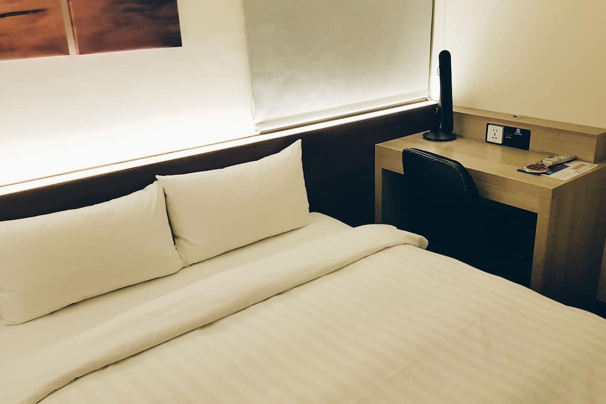 A hotel room with a bed, a desk with a chair, and a window with blinds.