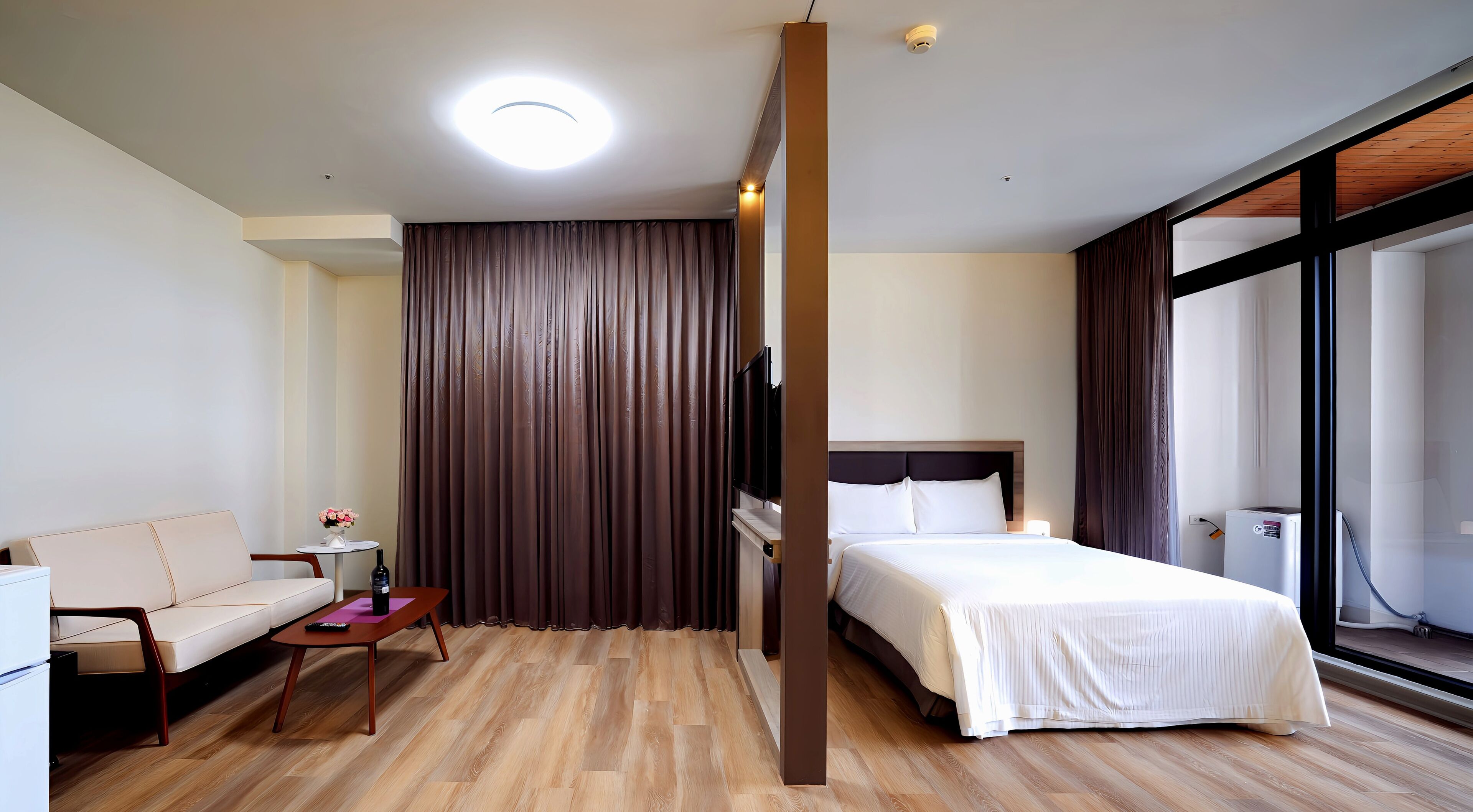 A hotel room with a bed, a sofa, a small table with a vase, and a large window.