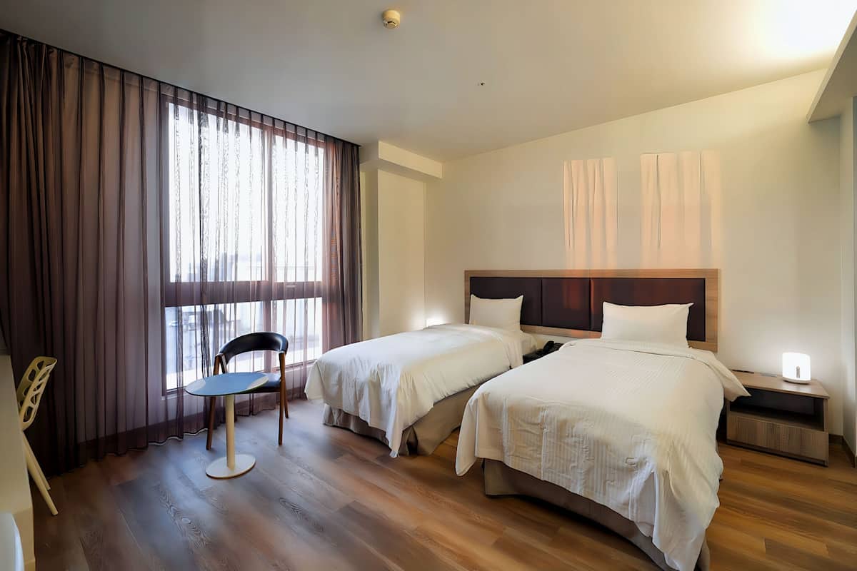 A hotel room with two beds, a chair, a bedside table with a lamp, and a window with curtains.