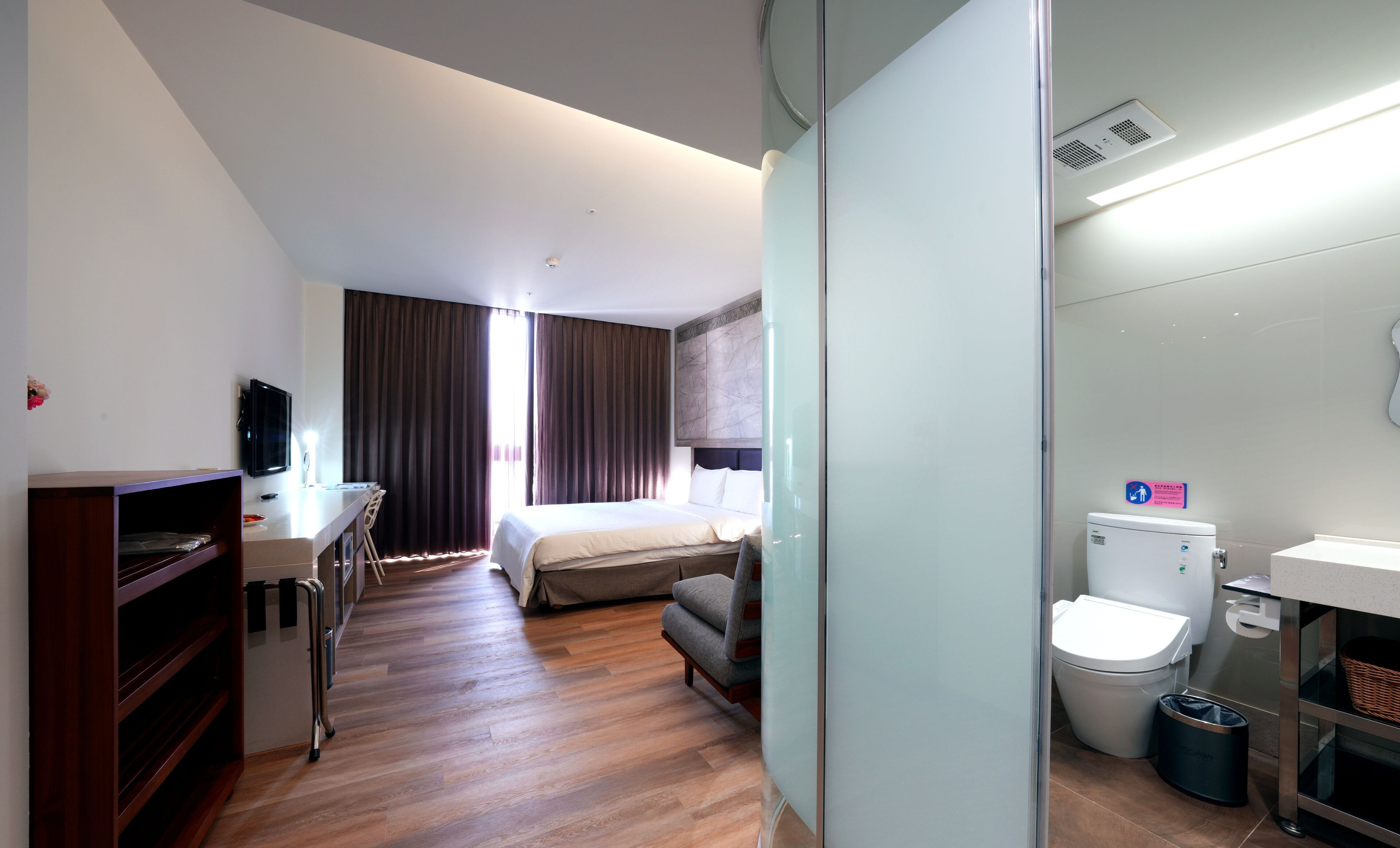 Camera d'albergo con un letto, una scrivania con sedia, una televisione, un bagno con il water e il lavandino, e un cestino.