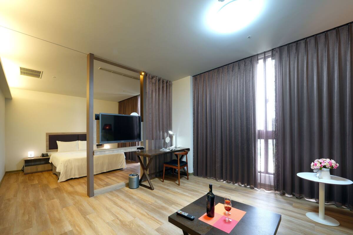 A hotel room with a bed, TV, desk, chair, and a small table with a bottle and glass.