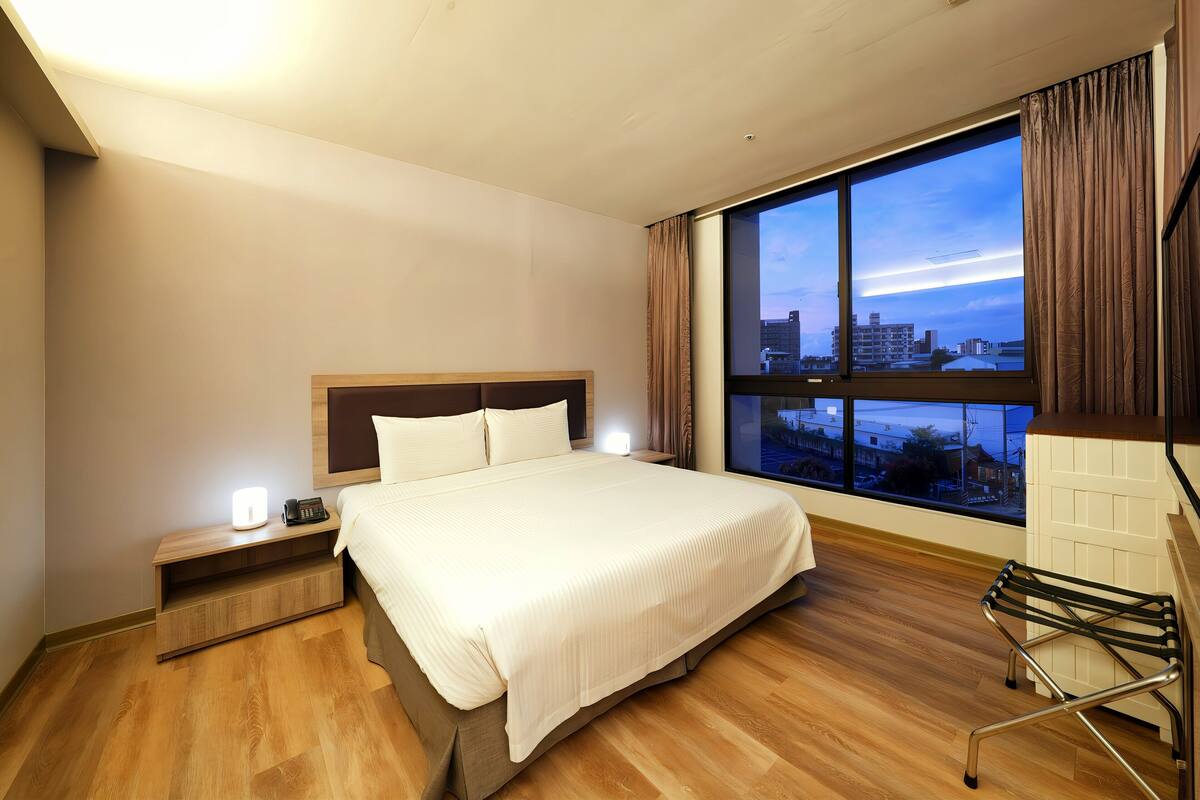 A hotel room with a large bed, a nightstand with a lamp, a chair, and a view of the city through a large window.