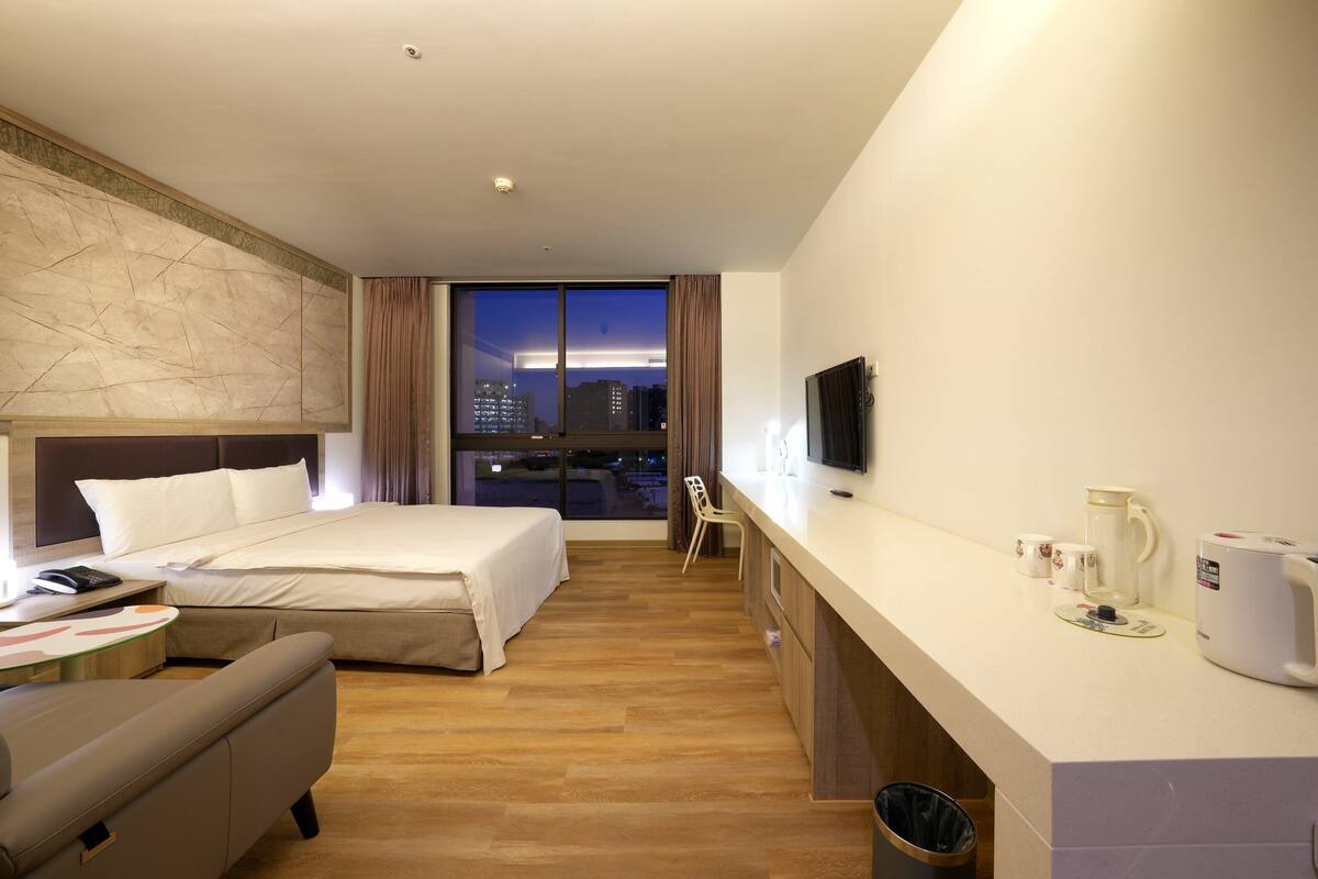 A hotel room with a bed, a desk, a chair, a TV, and a view of the city at night.