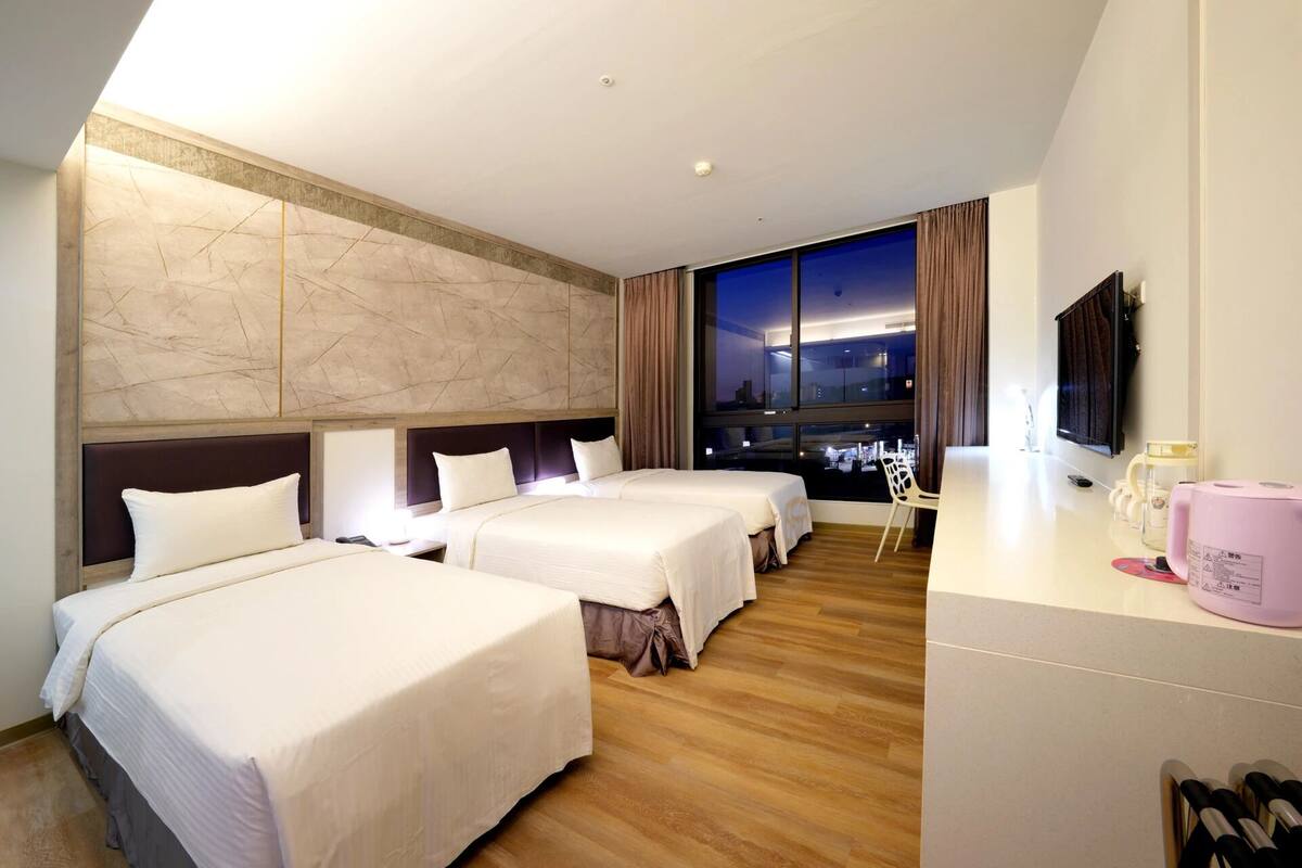 A hotel room with two beds, a desk with a pink water dispenser, and a large window with a city view.