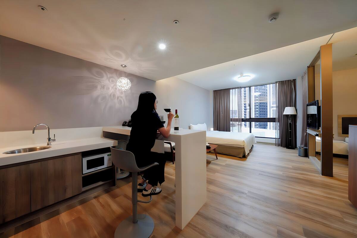 A modern hotel room with a kitchenette, a dining area, a bed, and a large window with curtains.