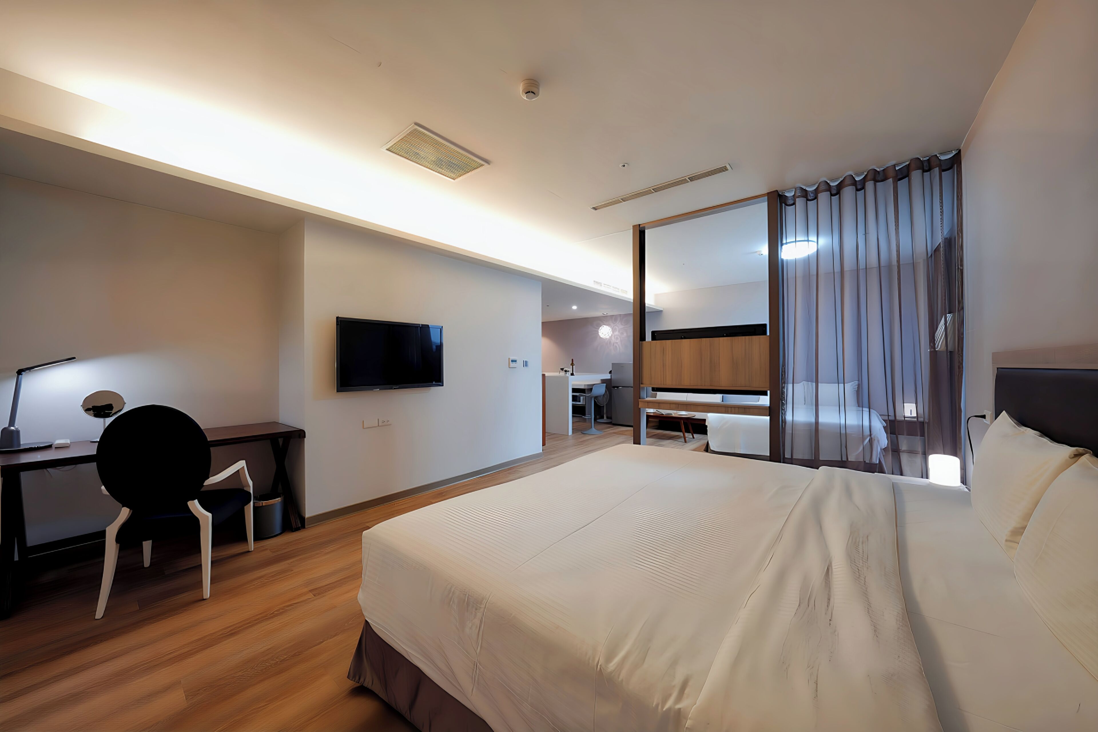 A hotel room with a large bed, a desk with a chair, a TV, and a bathroom visible through a glass partition.