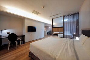 A hotel room with a large bed, a desk with a chair, a TV, and a bathroom visible through a glass partition.