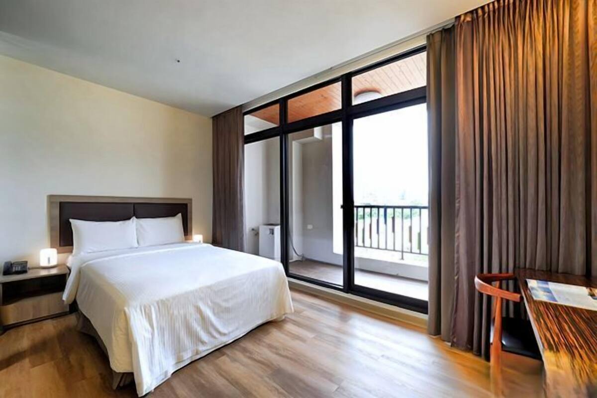 A hotel room with a large bed, a nightstand, a table, and a balcony with curtains.