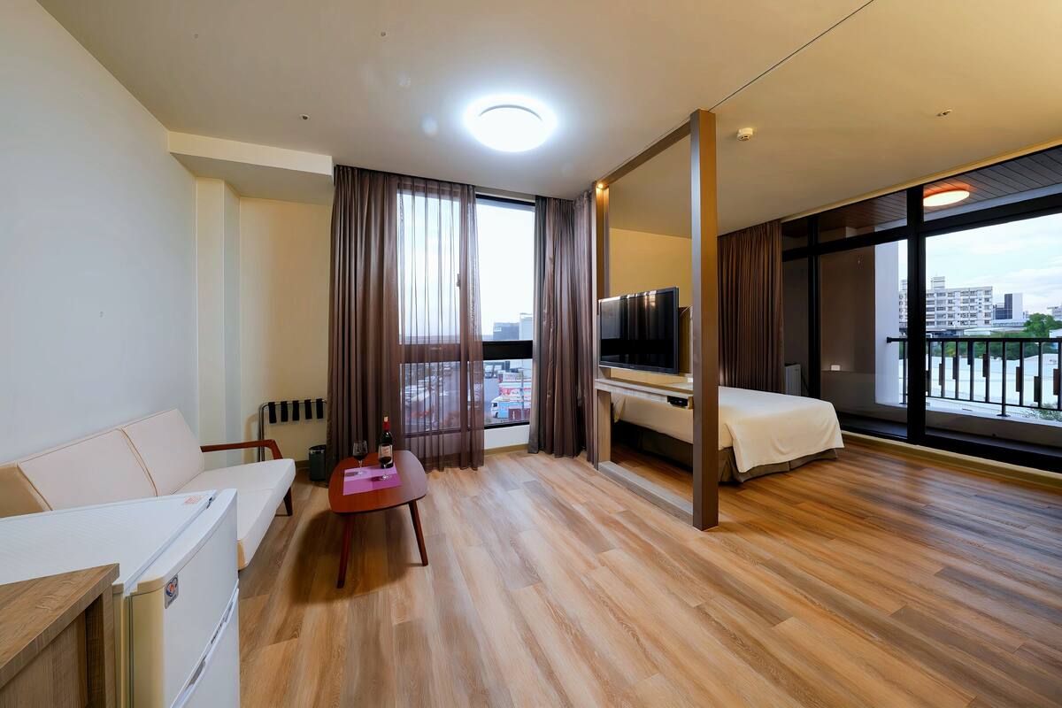 A modern hotel room with a wooden floor, a bed, a TV, a sofa, and a balcony with a city view.