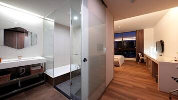 Una habitación de hotel moderna con ducha de vidrio, un amplio ventanal con vistas a la ciudad, una zona de estar independiente con televisor y suelo de madera.