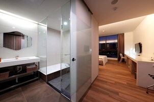 Una habitación de hotel moderna con ducha de vidrio, un amplio ventanal con vistas a la ciudad, una zona de estar independiente con televisor y suelo de madera.