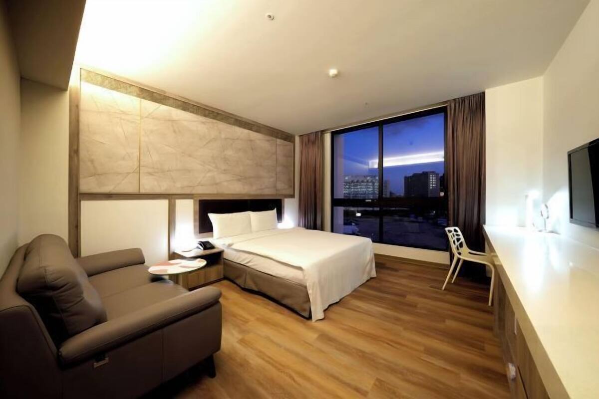 A modern hotel room with a bed, a sofa, a desk, and a large window with a city view.