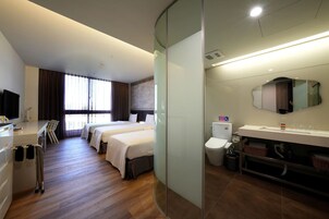 Habitación de hotel con dos camas, un escritorio con silla, un ventanal con cortinas, y un baño con inodoro, lavabo y espejo.