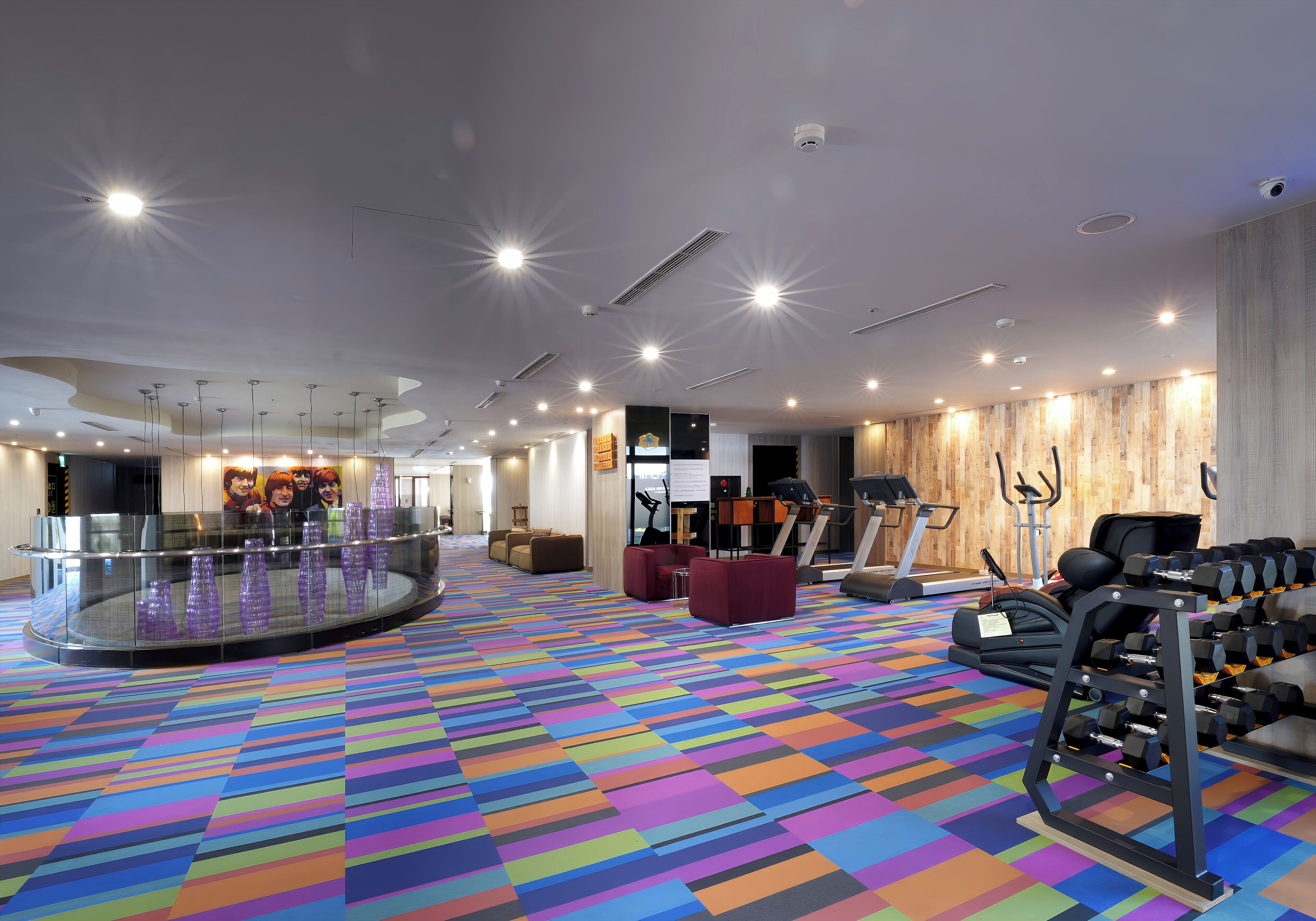 A spacious hotel lobby with a colorful carpet, a glass railing, fitness equipment, and a seating area.