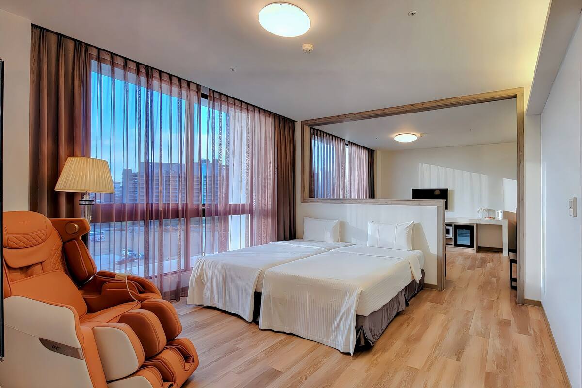 A hotel room with a large bed, a massage chair, a desk, and a view of the city.