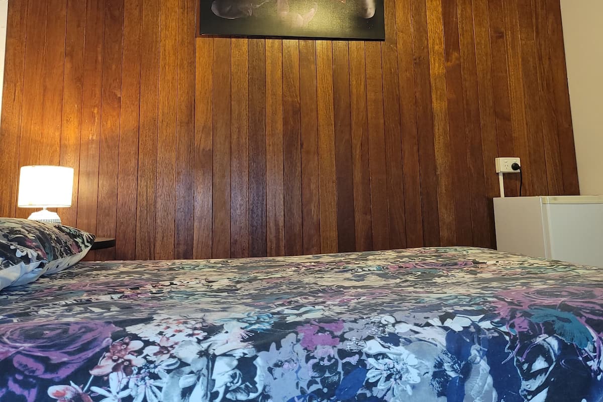 Ein Schlafzimmer mit einer Wandpaneele aus Holz, einem Deckenventilator, einem Bett mit Blumendekor und einer an der Wand befestigten Lampe.