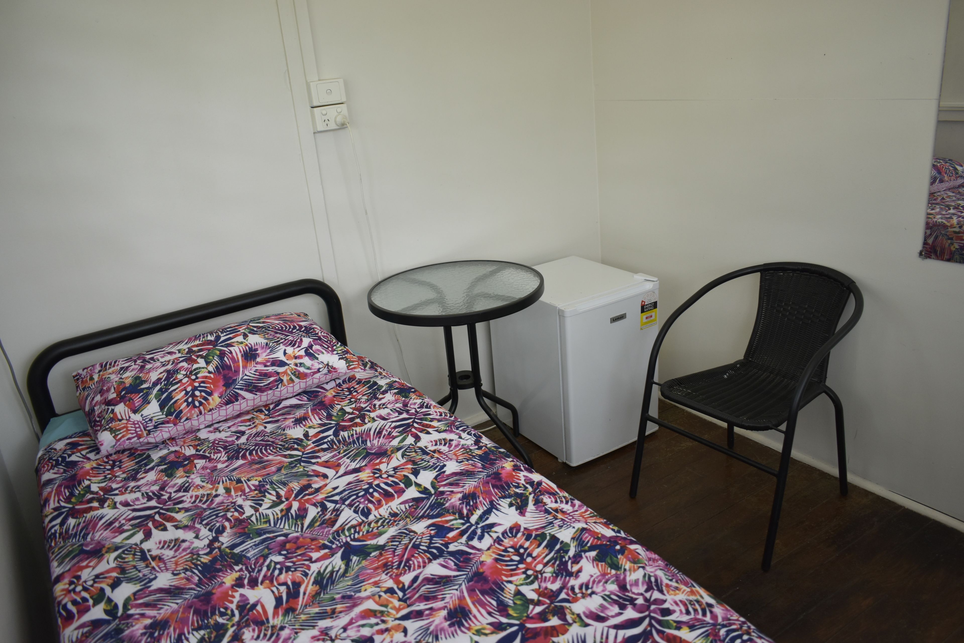 A small room with a bed, a table, a chair, and a white refrigerator.