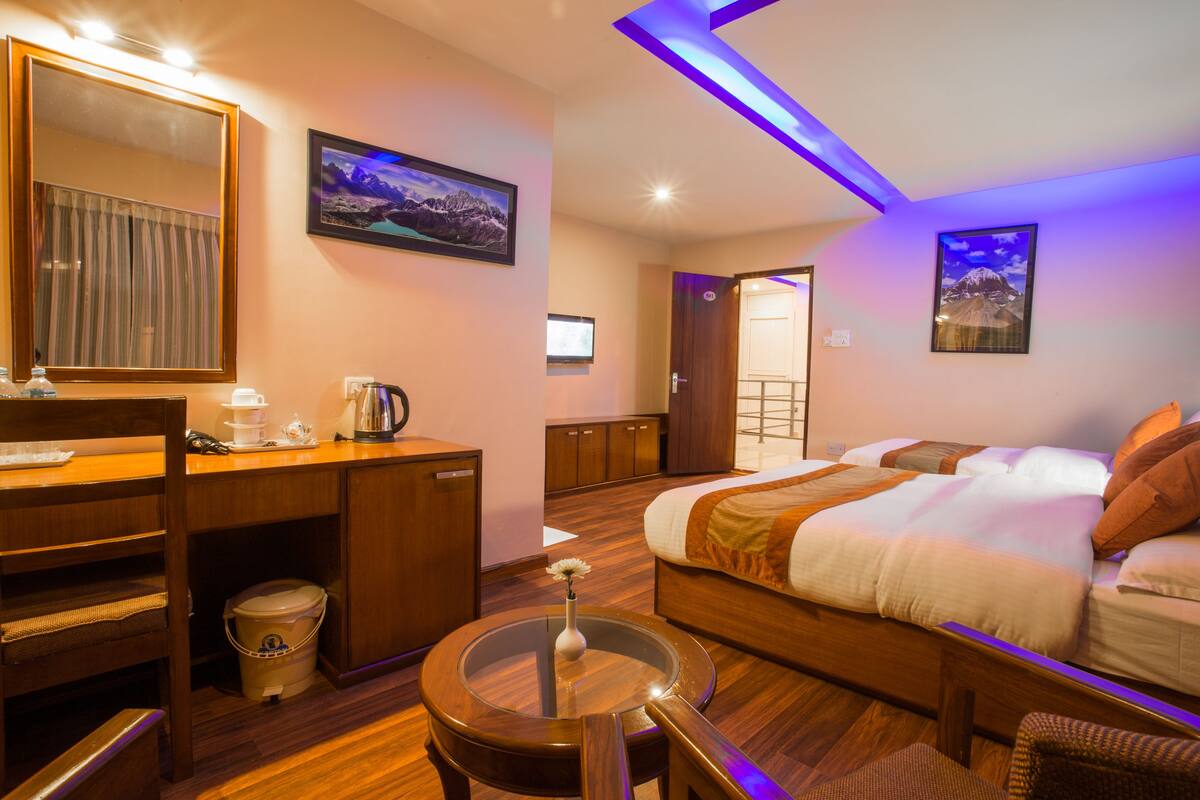A hotel room with a bed, a desk, a chair, a small table with a vase, a bucket, a mirror, and a TV.