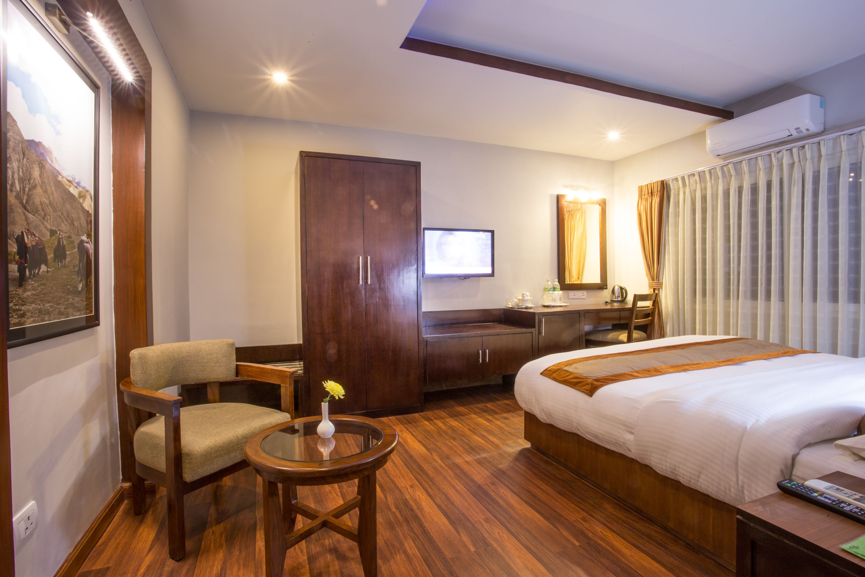 A hotel room with a bed, a desk, a chair, a small table with a vase, and a television.