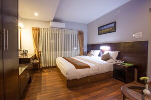 A hotel room with a wooden bed, a desk with a chair, an air conditioner, and a painting on the wall.