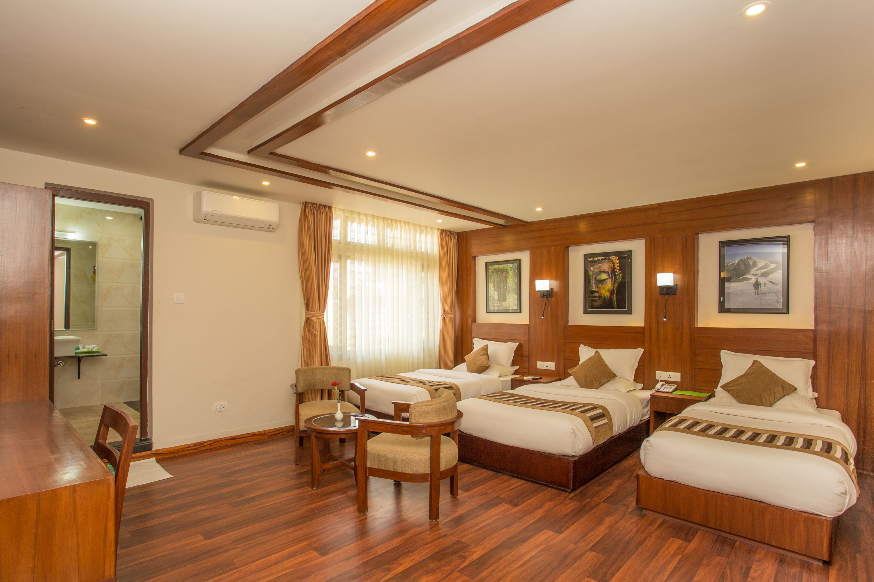 Habitación de hotel con dos camas, suelo de madera, zona de estar con una silla y una mesa, y baño visible a través de una puerta abierta.