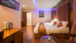 A modern hotel room with a large bed, a bedside table with a kettle, a glass table with a vase, and a view of a mountain through a window. - Aryatara Kathmandu Hotel (Kathmandu)