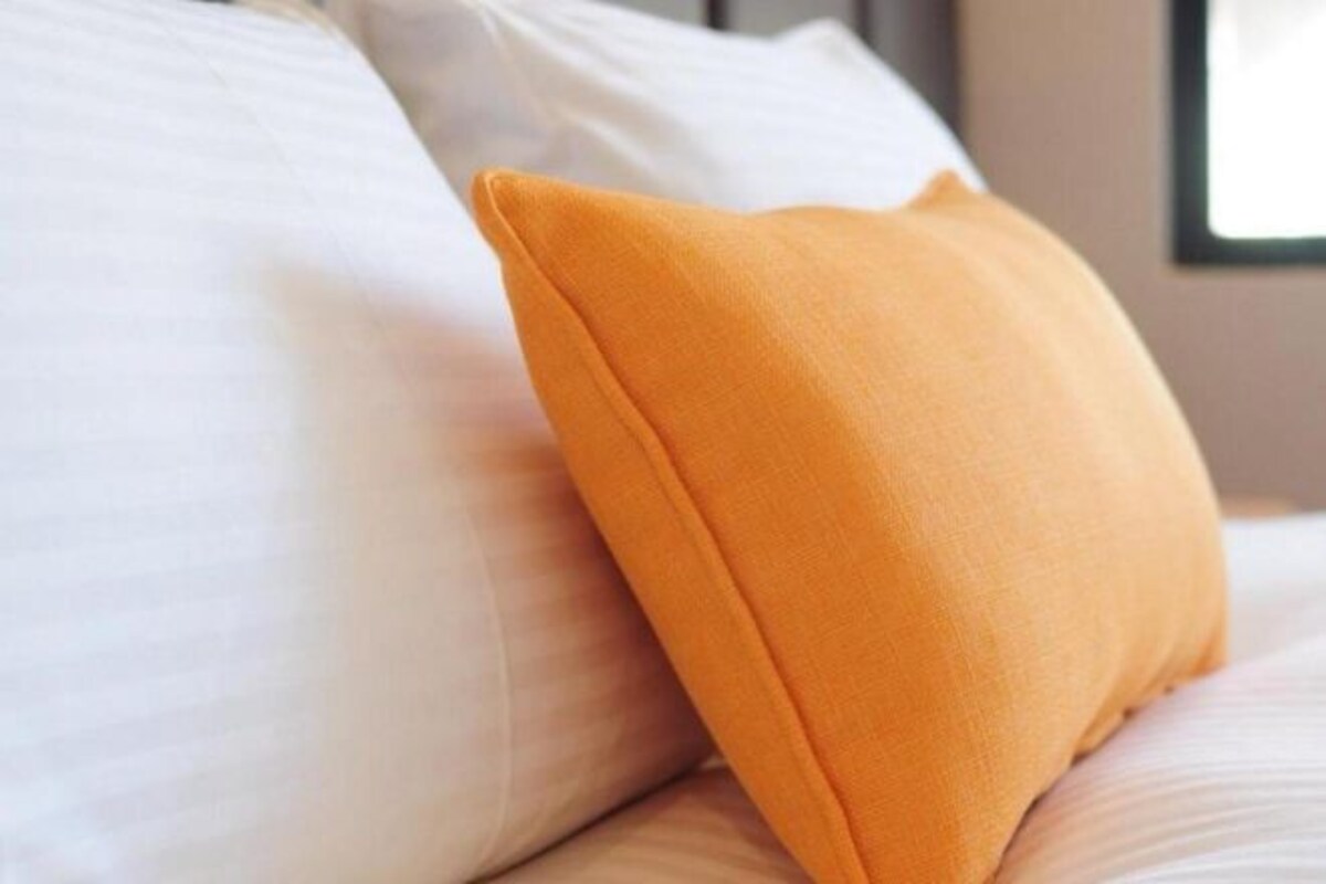 A bed with white bedding and a single orange pillow.