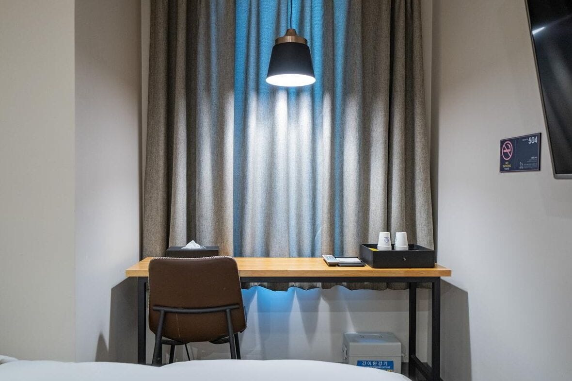 Habitación de hotel con escritorio, silla y ventana con cortinas. Hay un cartel en la pared y un televisor montado.
