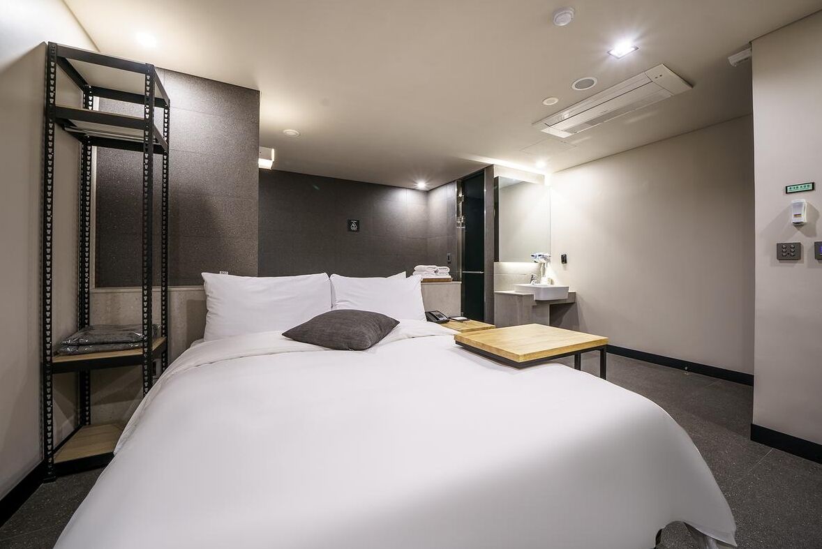 Hotellihuone, jossa on sänky, yöpöytä, peili, hylly ja seinälle asennettu ilmastointilaite.