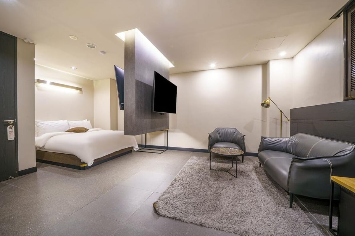 A modern hotel room with a bed, a sofa, a TV mounted on a wall, and a small table.