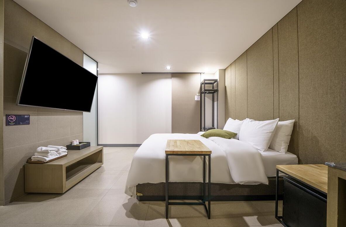 A modern hotel room with a large bed, a flat-screen TV mounted on the wall, a small wooden table, and a bench.