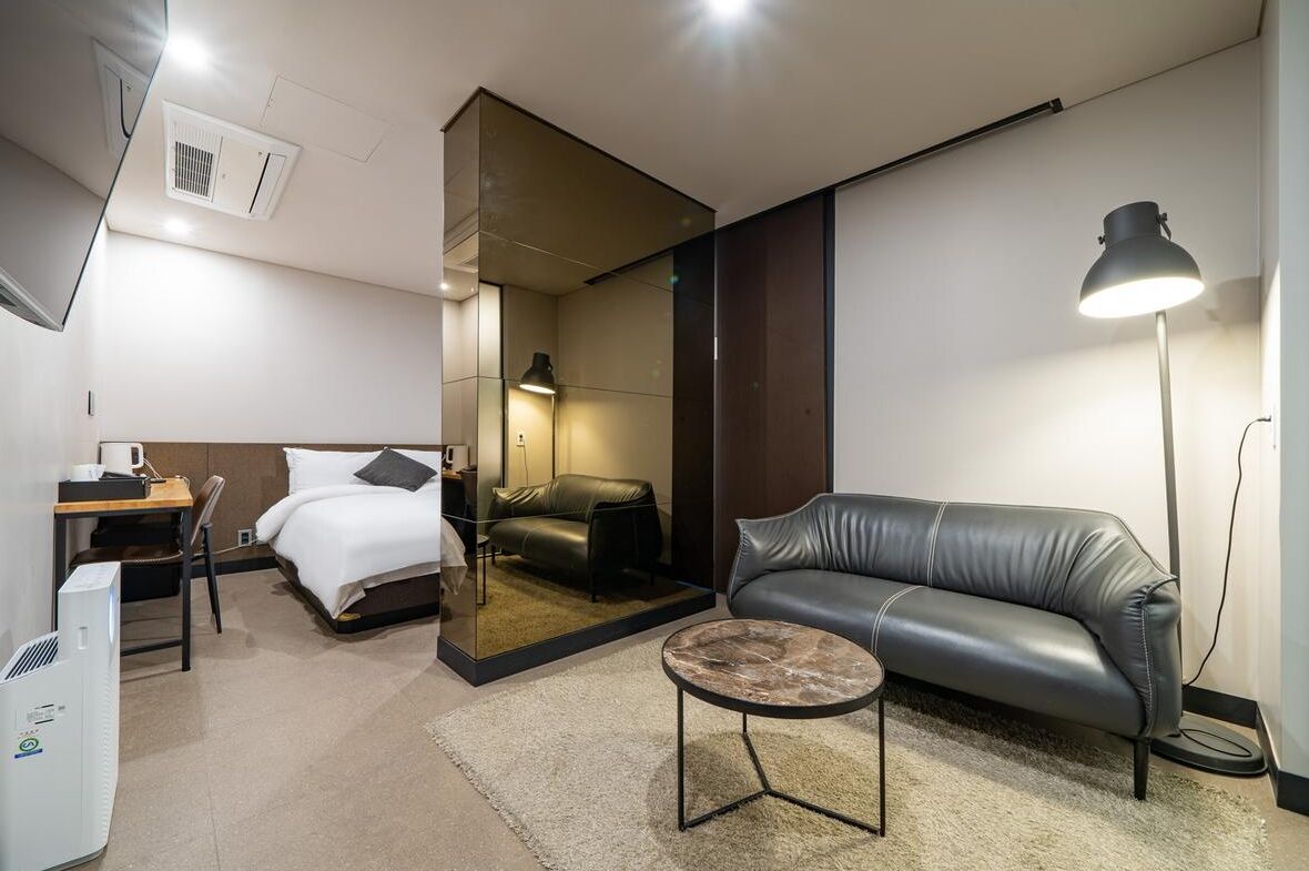 A modern hotel room with a bed, a leather sofa, a small round table, a desk with a chair, and a standing lamp.