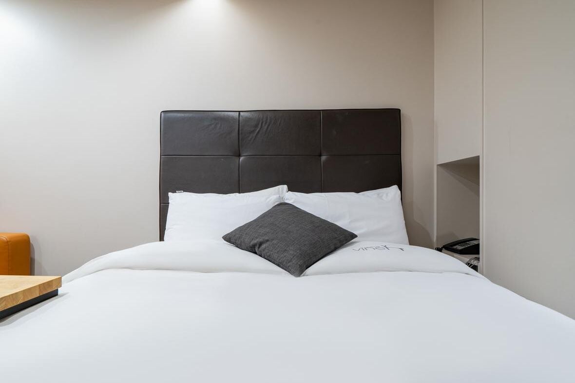 Cama bem arrumada com cabeceira preta, roupa de cama branca e um único travesseiro cinza.