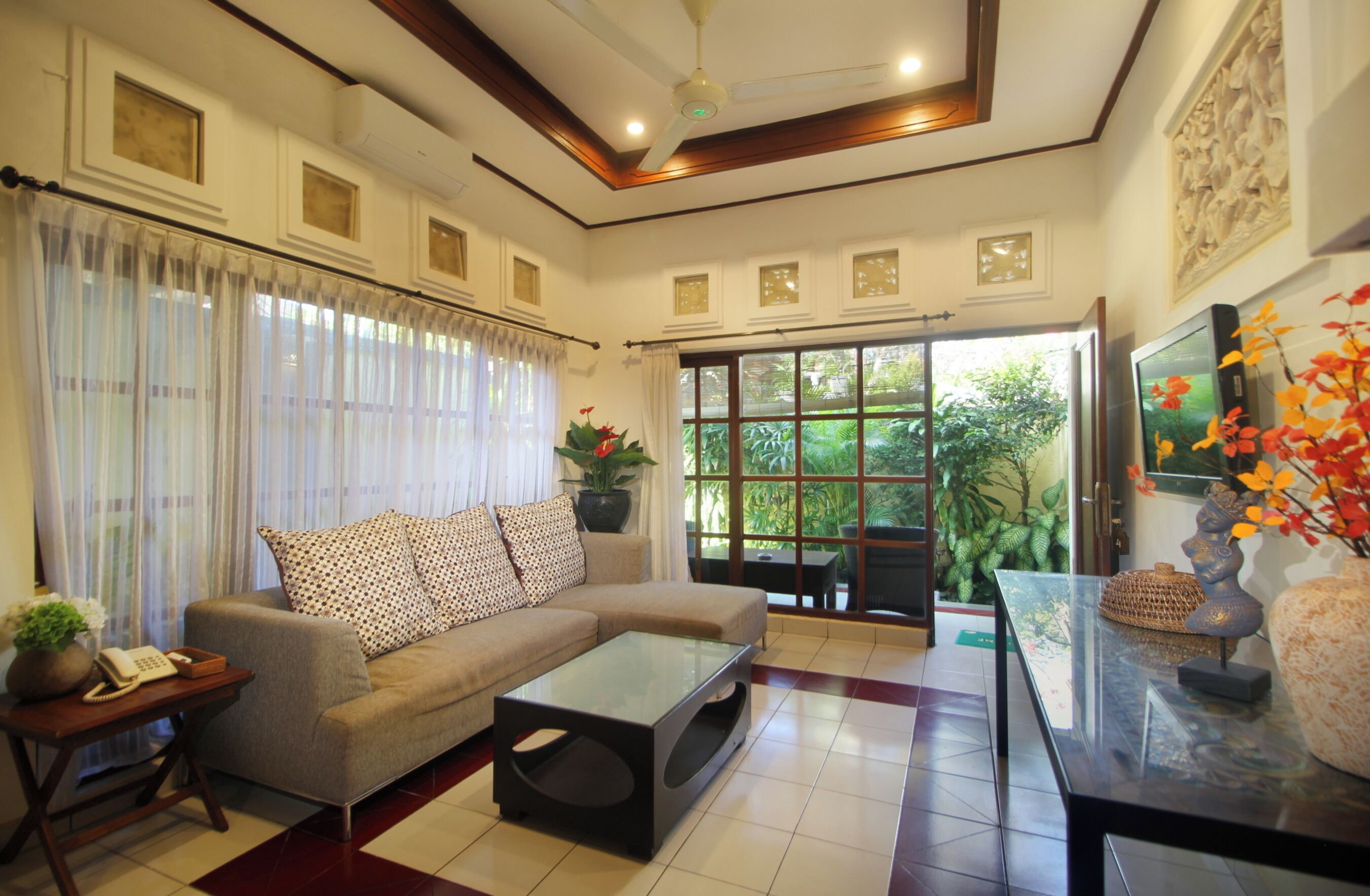 A living room with a sofa, coffee table, and a television. There is a window with curtains and a view of greenery outside.