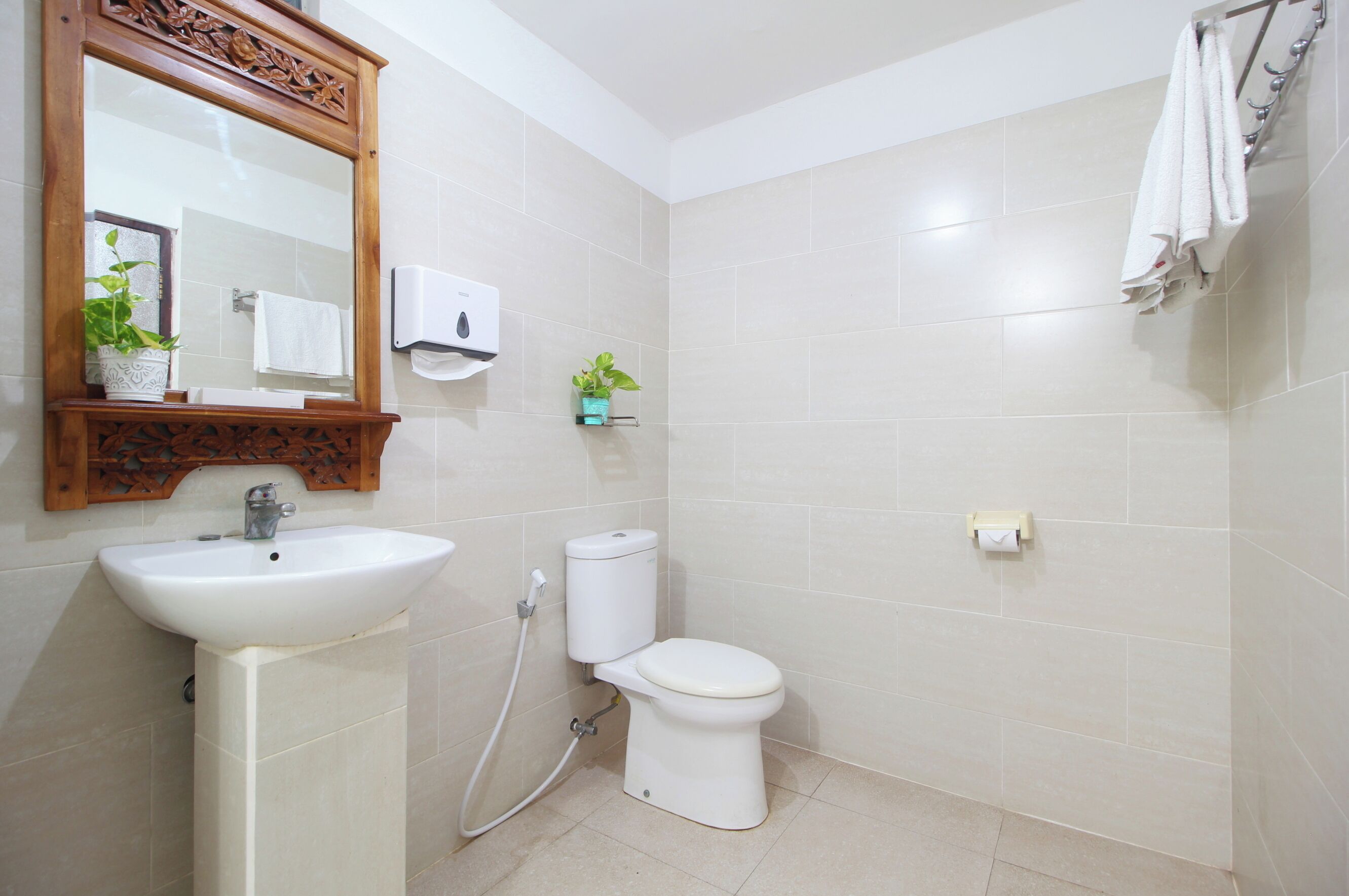A bathroom with a sink, toilet, and mirror. There are two potted plants, a towel rack, and a soap dispenser.