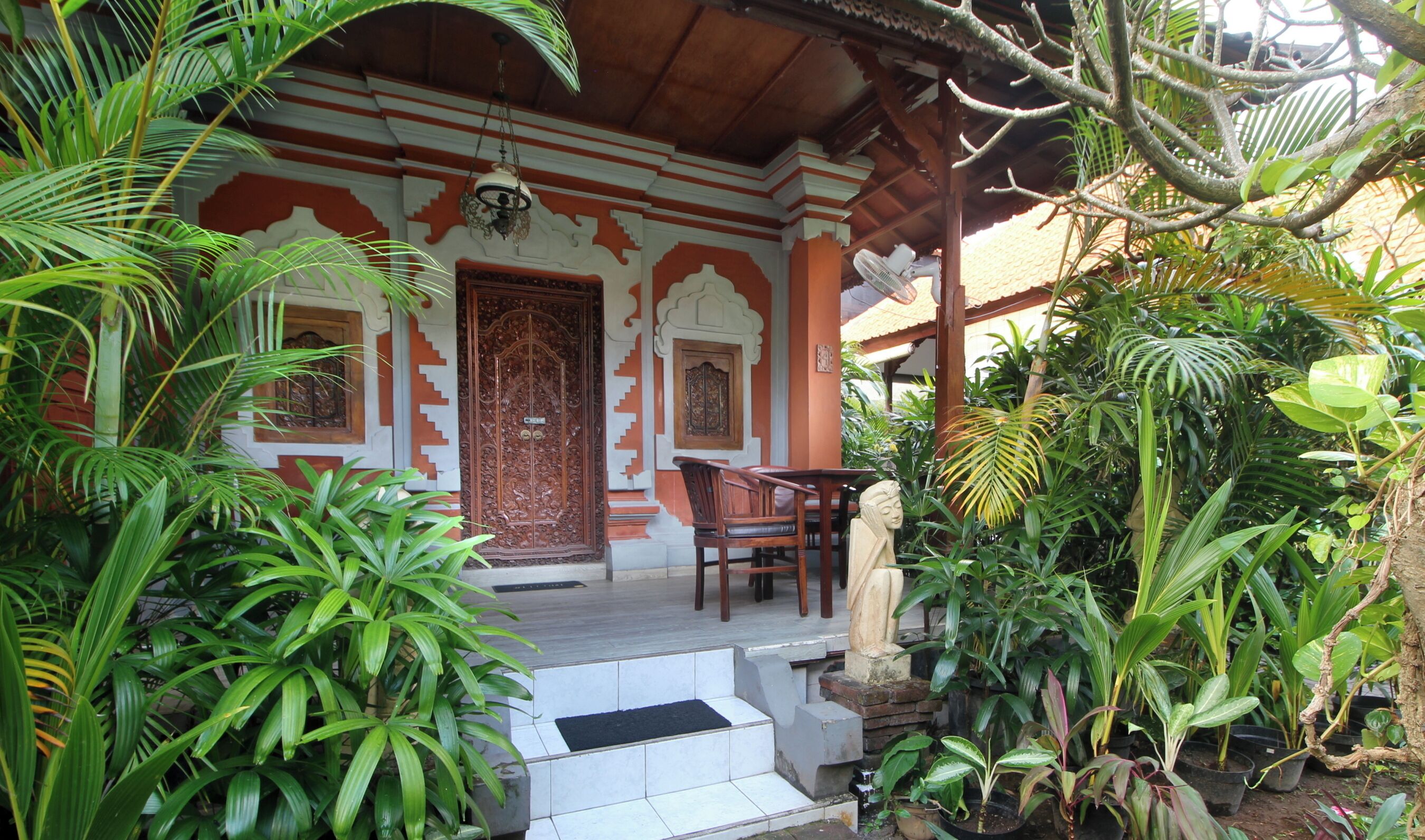 A traditional building with a wooden door, a porch with chairs, and lush tropical plants.