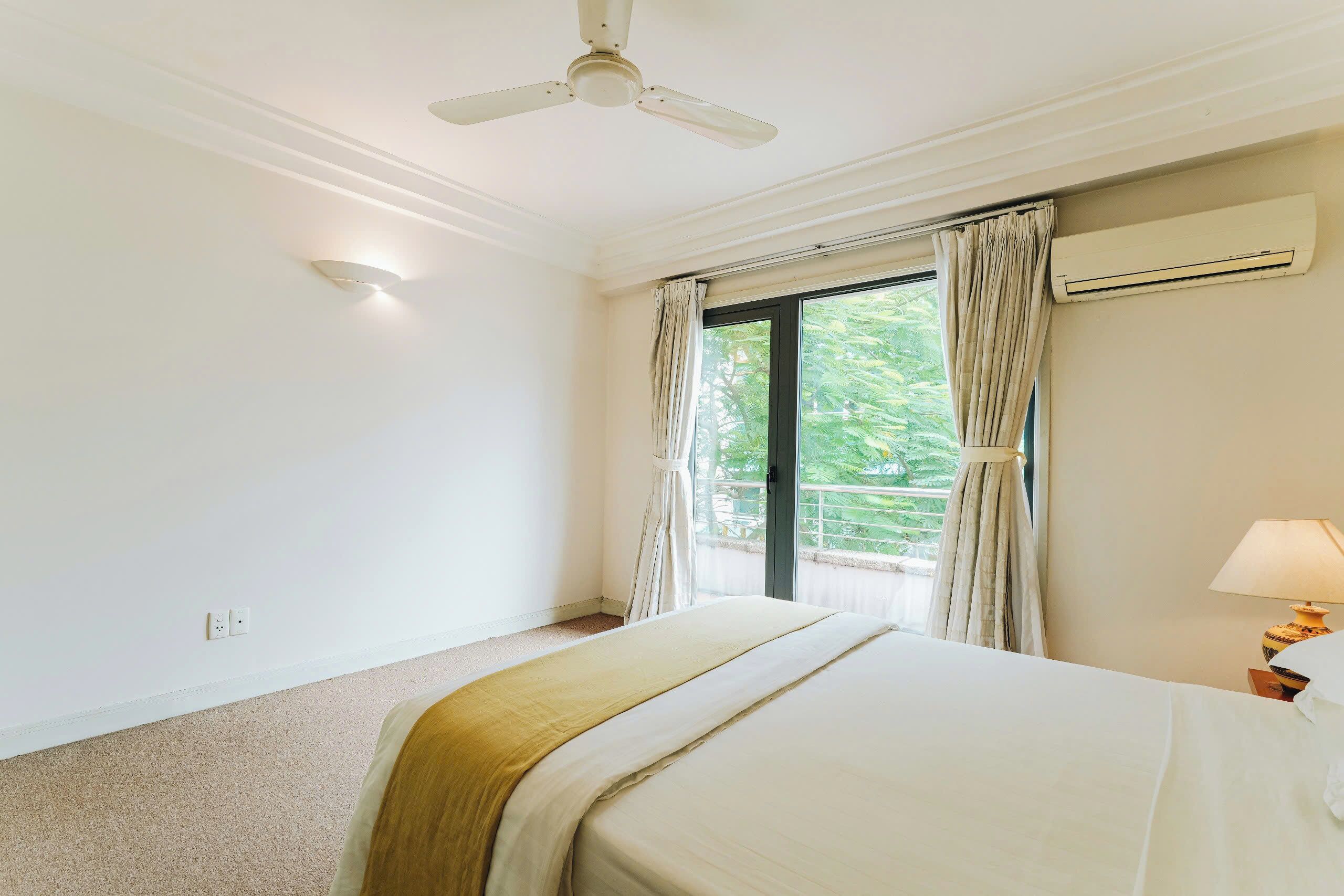 A hotel room with a bed, ceiling fan, air conditioning unit, window with curtains, and a nightstand with a lamp.