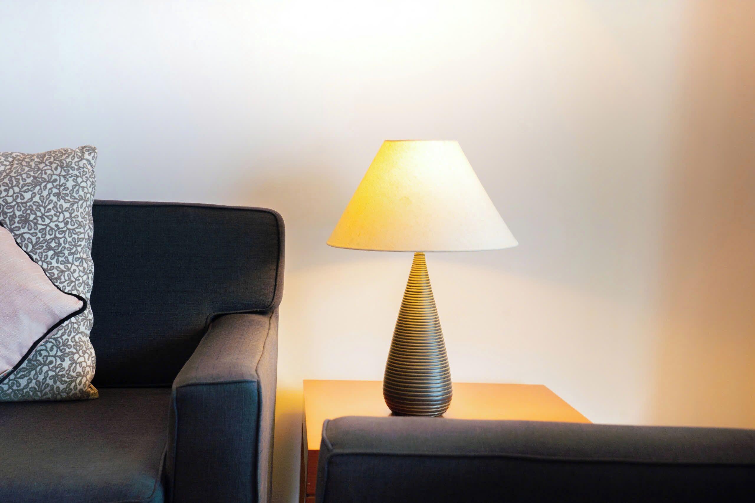A modern room with a dark sofa, a patterned pillow, and a conical table lamp on a bedside table.