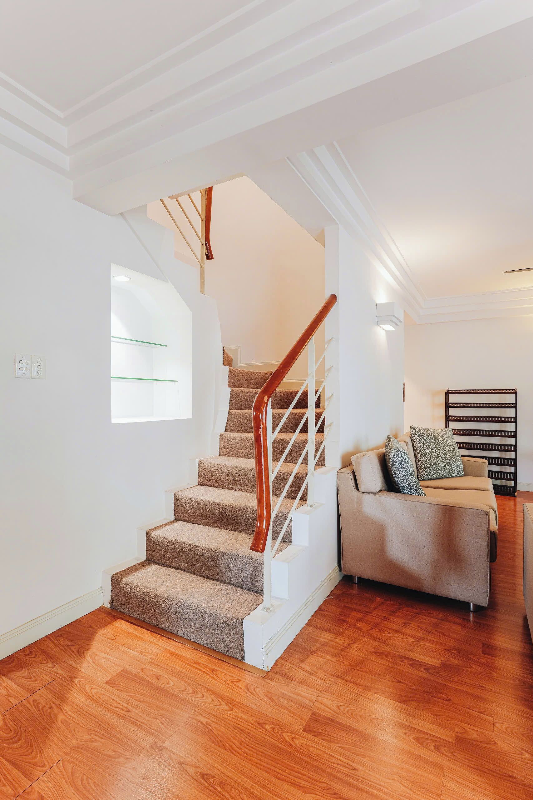 A staircase with wooden steps and a white handrail, leading to a landing area with a beige sofa and a window with a green frame.