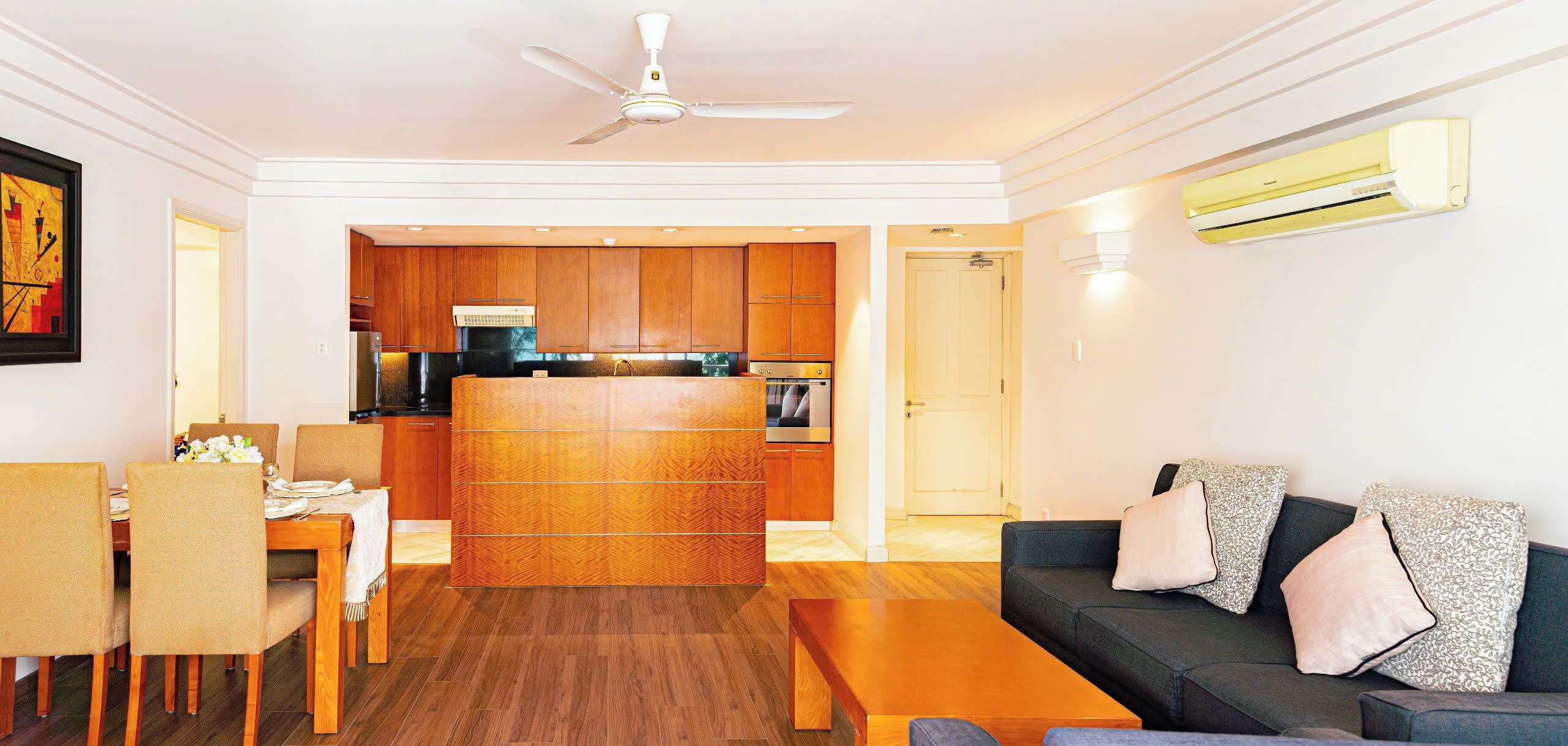 A modern living room with a sofa, coffee table, and dining area. There is a kitchen in the background with wooden cabinets and a refrigerator.