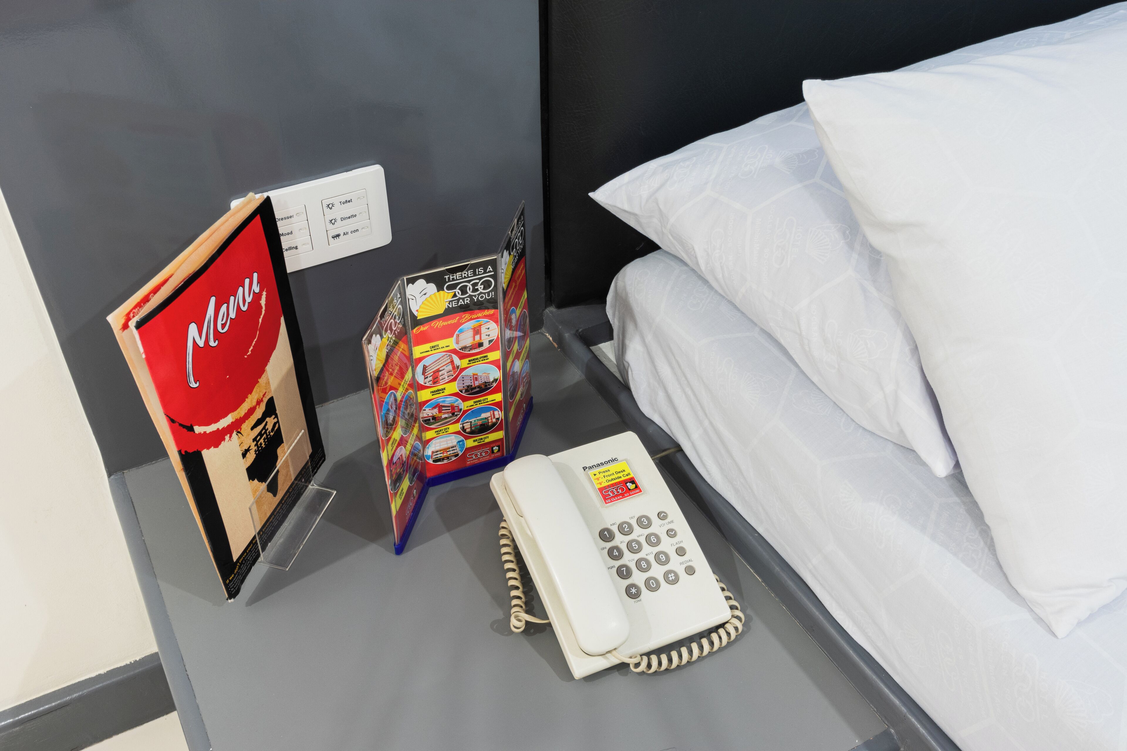 A hotel room with a bed, white pillows, a bedside table with a Panasonic phone, a menu stand, and a colorful brochure.
