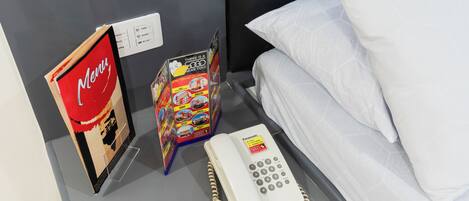 A hotel room with a bed, white pillows, a bedside table with a Panasonic phone, a menu stand, and a colorful brochure.