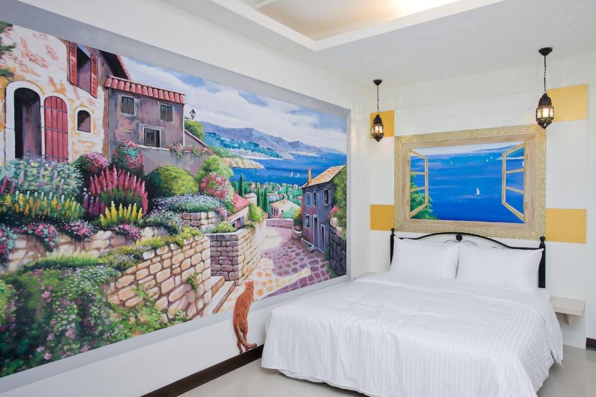 A bedroom with a mural of a village scene, a bed with white bedding, and a view of the sea through a window.