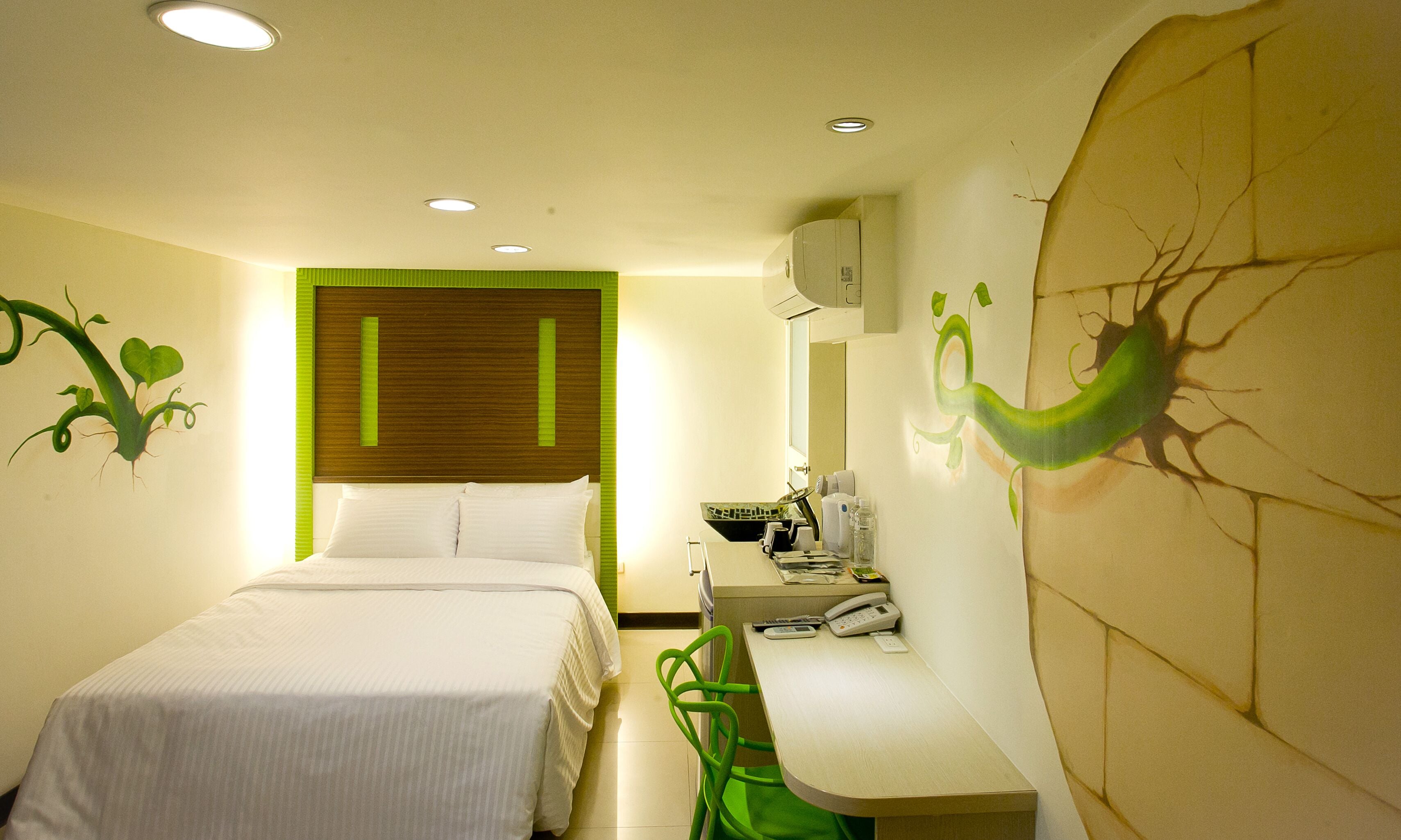 Quarto de hotel com cama, escrivaninha e cadeira. Há uma janela com persianas, parede com um desenho de planta e ar condicionado na parede.