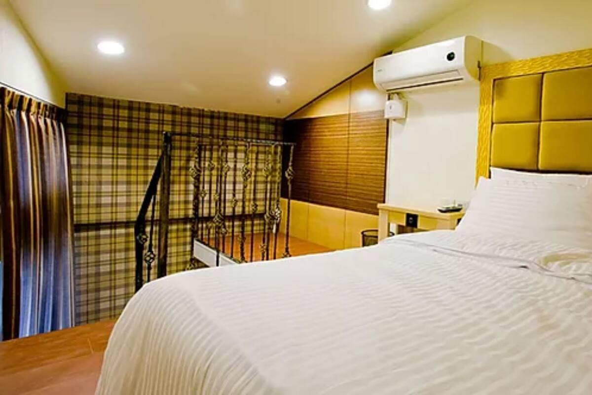 A hotel room with a bed, a nightstand, a window with curtains, and an air conditioning unit.