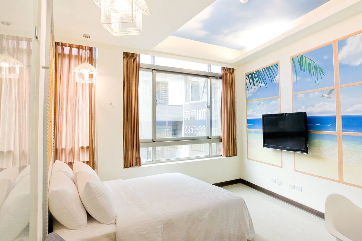 A hotel room with a bed, a TV mounted on the wall, a window with curtains, and a beach mural.