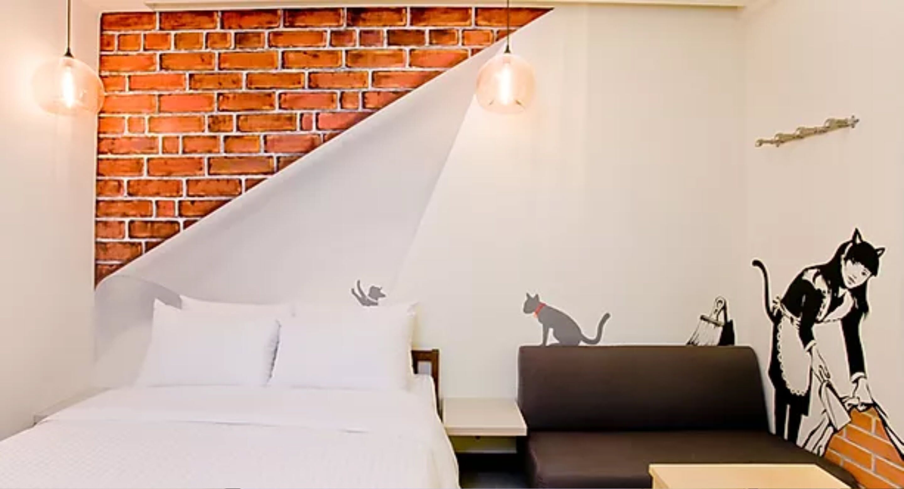 Une pièce comprenant un lit, un canapé, un mur en briques et une fresque représentant un chat et une personne.