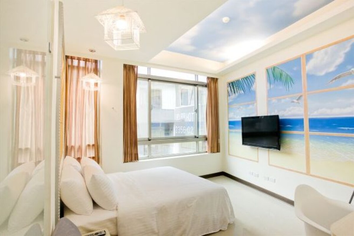 A hotel room with a bed, a TV mounted on the wall, a window with curtains, and a beach mural on the wall.